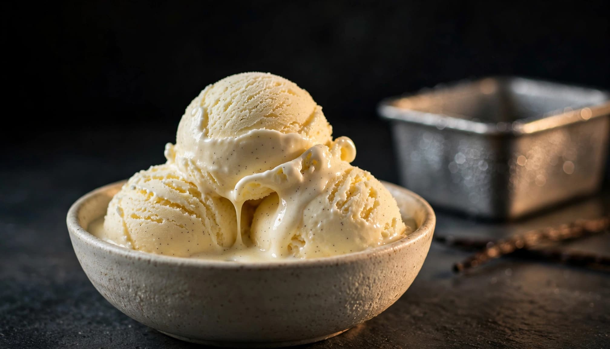 Soft scoops of vanilla ice cream in a bowl beside a metal pan and vanilla bean.
